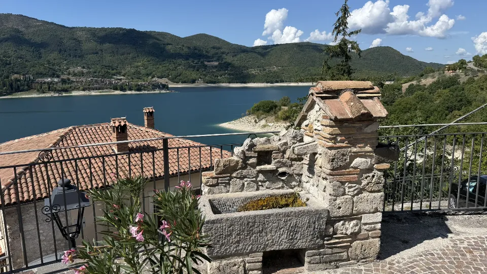 Antica fontana in pietra in un borgo affacciato sul Lago del Turano, con tetti in cotto, montagne verdi e il lago sullo sfondo.
