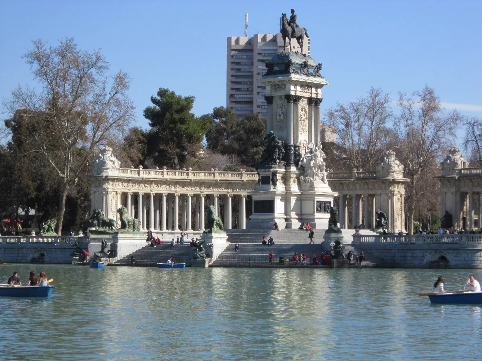 Vista del monumento ad Alfonso XII nel Parco del Retiro a Madrid, con il colonnato monumentale, la statua equestre e le barche sul lago, sotto un cielo azzurro.